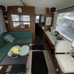 Photo de l'intérieur du bateau depuis la porte d'entrée. On voit à gauche une banquette avec une table rectangulaire grise, une fleur jaune et deux sets de table et à droite le poste de pilotage intérieur du bateau.