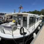 Vue de côté du bateau avec sa coque blanche et noire. On voit le pont avant avec une table ronde de jardin et deux chaises. Un faux bouquet de fleurs jaune est disposé sur la table.