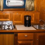 Image de la cuisine du bateau tout en bois. On y voit à gauche le réfrigérateur et la plaque de cuisson au gaz et à droite l'évier ainsi qu'une cafetière.