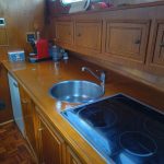 Image de la cuisine du bateau avec placards et plan de travail en bois. On voit sur la photo à droite la plaque de cuisson vitrocéramique, un évier et des accessoires de cuisine (cafetière, boîte à sucre) tout à gauche.