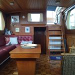 Photo de la pièce à vivre du bateau. On y voit un petit escalier en bois permettant d'accéder à la pièce. A gauche, une banquette en cuir bordeaux avec une table basse en bois et à droite un fauteuil en osier.